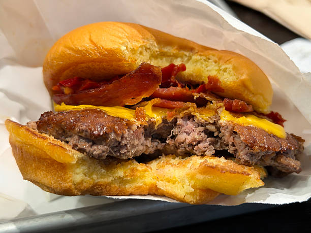 Stock photo shows a close up of a man taking a bite while eating a cheeseburger beef burger hamburger layered with melted cheese and crispy bacon inside a fresh bread roll. Juicy fast food takeaway meal showcasing indulgent street food flavors, appetite, satisfaction