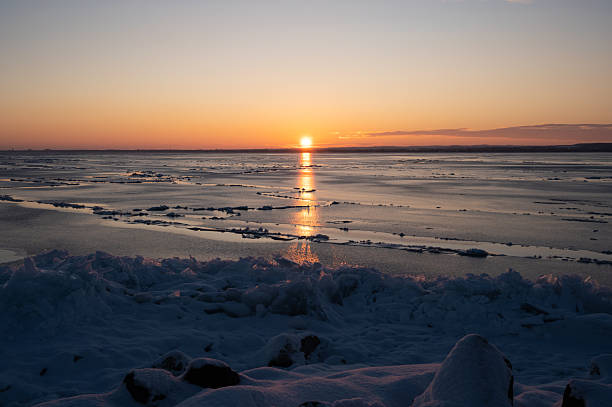 A serene winter sunrise over the Lake Balaton, with golden light reflecting across the frozen surface and broken ice formations. The calm horizon and warm colors contrast beautifully with the cold, icy foreground.