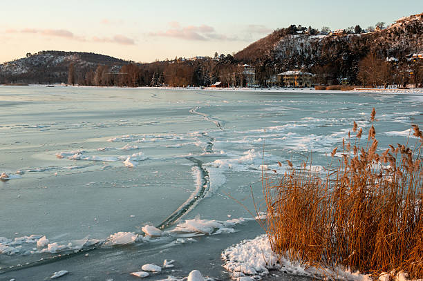 The frozen Lake Balaton covered with cracked ice stretches across a peaceful winter landscape. Snowy hills, bare trees, and warm sunset light create a calm and atmospheric seasonal scene.