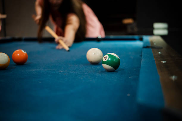 Selective focus photograph of billiard balls spread across a blue pool table, with a player aiming a cue stick in the background. Concept of strategy, precision, skill, and indoor leisure activity. Ideal for themes related to billiards, pool game, competition, recreation, entertainment, and lifestyle sports.