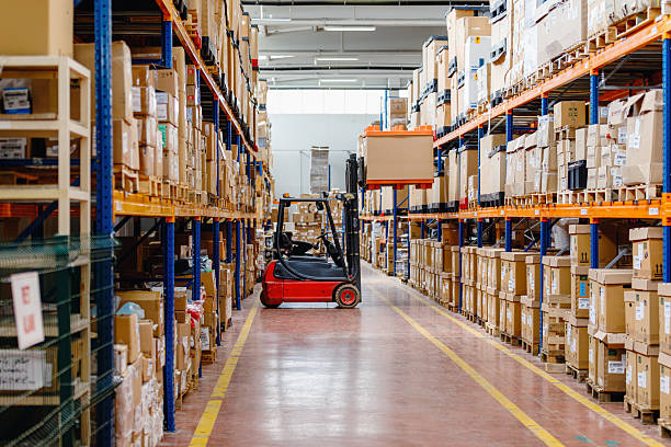 Wide view of a modern warehouse interior featuring a forklift parked in a long aisle between high rack storage shelves filled with cardboard boxes and pallets. The image represents advanced logistics operations, warehouse automation, inventory management, and smart supply chain infrastructure, highlighting technology driven efficiency, data based stock control, and artificial intelligence supported warehouse systems used in distribution, manufacturing, and e commerce fulfillment.