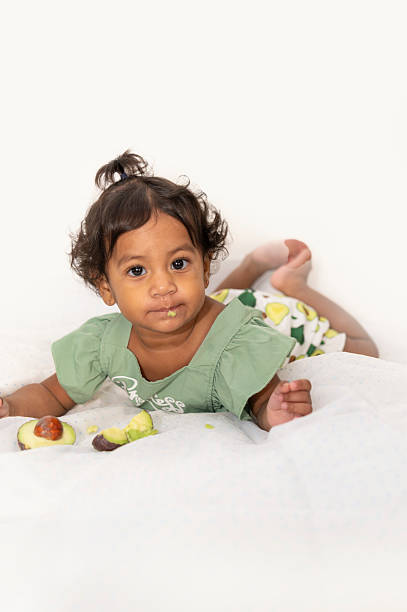 Adorable baby girl lying on tummy, discovering new flavors and textures with avocado during first meal