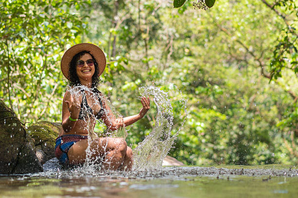 A mature woman is joyfully playing in a serene river during summer. She splashes water around, creating beautiful arcs as her hair flows, surrounded by lush greenery under a bright sky.