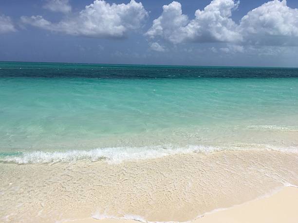 Gentle waves wash over white sandy beach under a partly cloudy sky. Turquoise ocean water stretches to the horizon. Calm tropical seascape with peaceful beach setting.