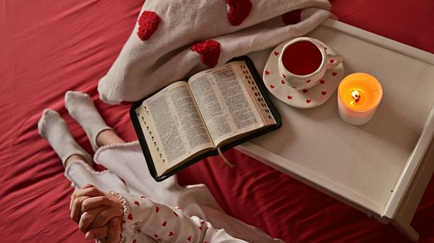 Close-up of a teenage girl reading a black book, with a rosary draped over her hand, suggesting a spiritual or religious context within a comfortable home setting.