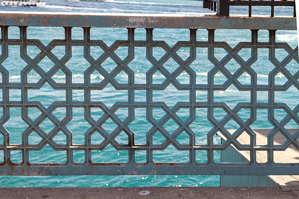 Close up view of an ornate metal railing featuring traditional geometric patterns on Galata Bridge with turquoise blue sea in the background, photographed in Istanbul, Turkiye, highlighting Ottoman inspired design, Islamic geometric motifs, weathered metal texture, coastal atmosphere, and iconic urban waterfront scenery suitable for travel, culture, architecture, texture, and abstract background concepts
