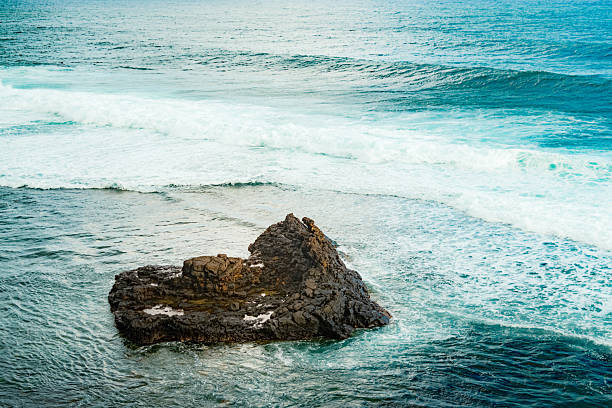 A heart-shaped rock rises from turquoise waters, showcasing oceanic power and serene beauty. Textured stone erosion patterns swirl with sea foam under an overcast sky; a unique geological marvel.