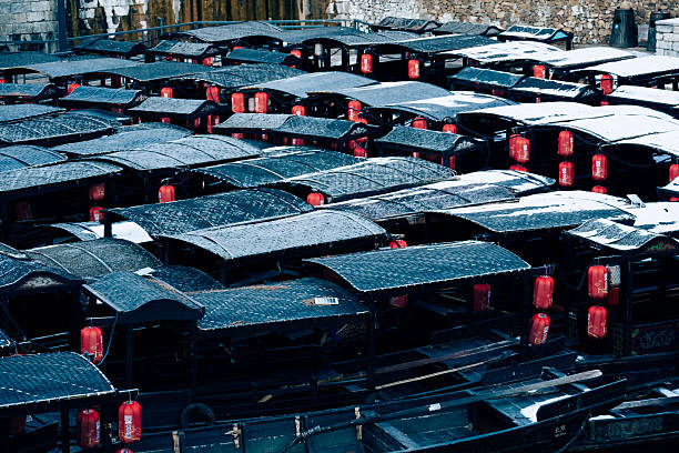 A row of traditional black canopied boats rests quietly at a snow-covered dock after a snowfall, their dark silhouettes contrasting starkly with the white, peaceful harbor scene.
