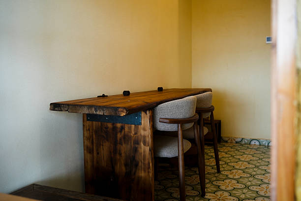 A cozy corner featuring a handcrafted wooden table made from reclaimed timber with visible grain and rough edges, supported by dark metal brackets. Two modern upholstered chairs with wooden frames are positioned beside it, set on a patterned tile floor with floral motifs. The warm yellow walls and soft lighting create a peaceful, inviting atmosphere, ideal for a home office, café, or rustic retreat.