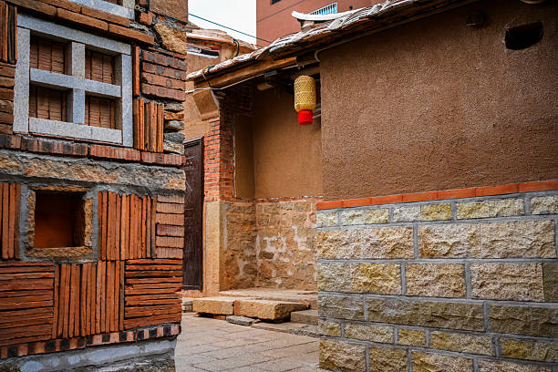 A narrow alleyway between traditional buildings featuring a mix of red brick, stone masonry, and earthen walls. The left wall displays decorative brick patterns and small window openings, while the right side has a smooth adobe-like surface with a tiled roof and a hanging bamboo lantern. The scene captures the quiet charm of a historic village, blending vernacular architecture with subtle cultural details. In the background, modern buildings hint at urban development, creating a contrast between old and new.
