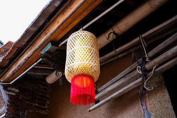 A traditional bamboo lantern hangs from the wooden eaves of an old building, its intricate lattice design glowing softly with warm light. The lantern features a vibrant red tassel at the bottom, adding a festive touch to the rustic scene. Surrounded by weathered wood beams, terracotta tiles, and earthen walls, this image captures the essence of Chinese folk craftsmanship and cultural heritage. Ideal for themes related to tradition, handmade art, festivals, or architectural details.