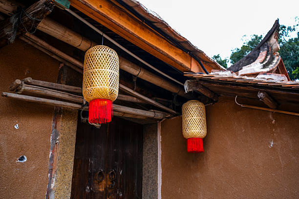 A traditional bamboo lantern hangs from the wooden eaves of an old building, its intricate lattice design glowing softly with warm light. The lantern features a vibrant red tassel at the bottom, adding a festive touch to the rustic scene. Surrounded by weathered wood beams, terracotta tiles, and earthen walls, this image captures the essence of Chinese folk craftsmanship and cultural heritage. Ideal for themes related to tradition, handmade art, festivals, or architectural details.