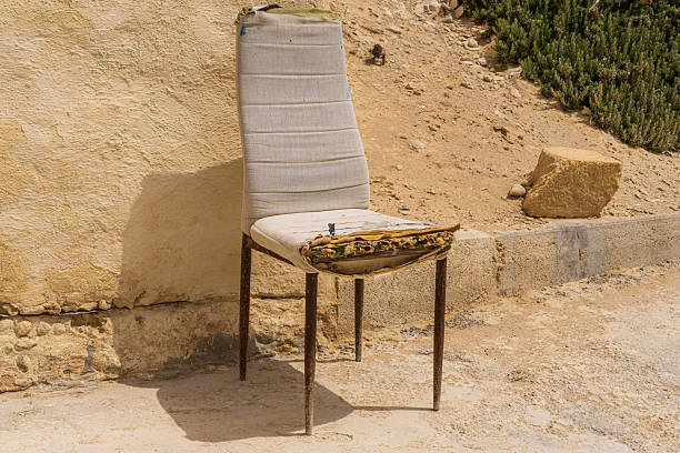 A damaged upholstered chair with exposed padding placed against a sunlit wall, conveying neglect, fragility, and quiet abandonment