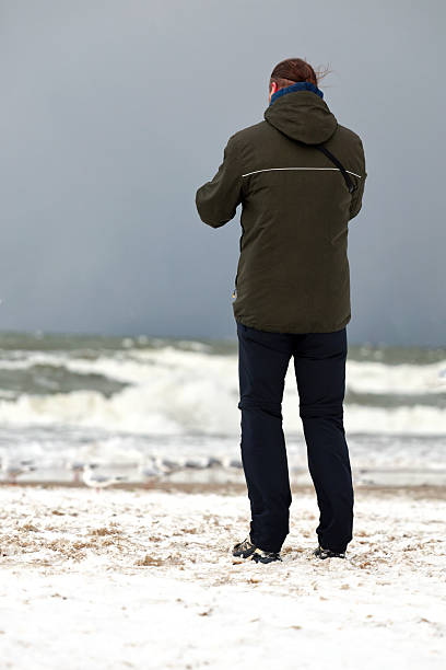 Person wearing a warm red coat and hat embracing a small dog while standing on a rugged shoreline, looking out at the calm blue ocean and distant cargo ships
