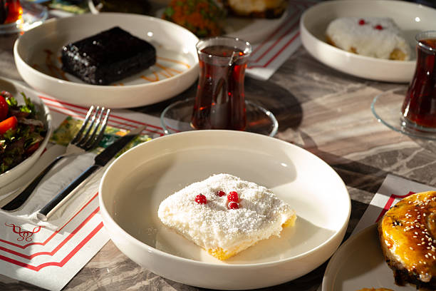 A high-quality close-up of a delicious traditional Turkish milk dessert topped with coconut and red berries. The scene is set on a rich dining table with blurred tea glasses and other side dishes in the background, illuminated by warm natural sunlight. This image captures a cozy and authentic family meal moment, ideal for culinary blogs, lifestyle magazines, and Mediterranean food culture content.