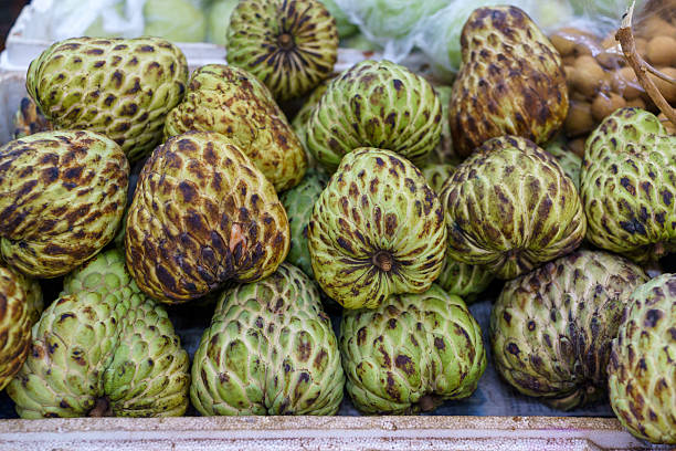 Close-up view of fresh sugar apples, also known as custard apples, stacked together at a local market. The tropical fruits show natural green skin with textured segments and ripening marks, commonly found in Southeast Asian markets and used in fresh consumption and desserts.