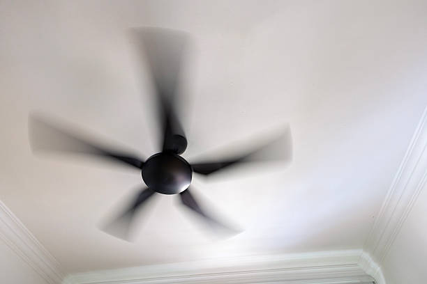 Low-angle view of a modern ceiling fan spinning on a white ceiling inside a residential interior. The motion blur emphasizes airflow, cooling, and everyday home comfort, representing household equipment and simple living
