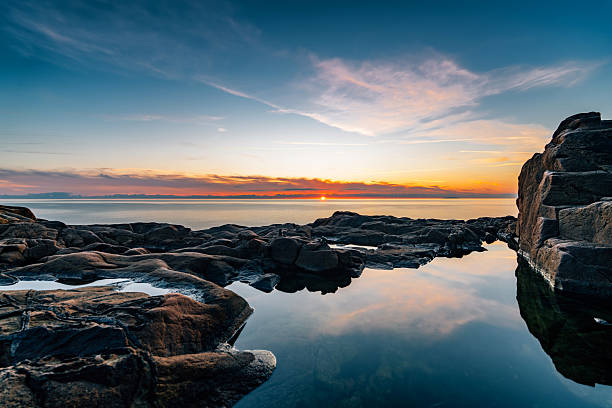 A breathtaking wide-angle view captures a vibrant sunset or sunrise over a rugged rocky coastline. The colorful sky, painted with hues of orange, blue, and pink, reflects beautifully in a calm tidal pool in the foreground. This tranquil natural landscape with a calm ocean stretching to the horizon creates a peaceful and scenic natural escape. Calafuria, Livorno, Tuscany, Italy.
