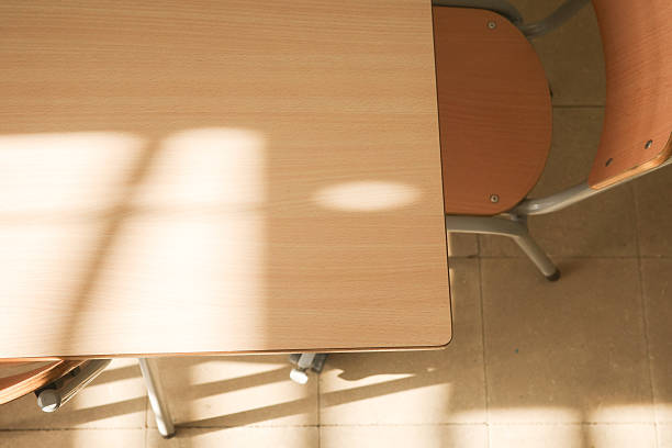 Top view of an empty school desk illuminated by natural sunlight creating soft window shadows. Minimal educational background representing classroom environment, learning space, school furniture, calm academic atmosphere, and education concepts. No people, copy space available.