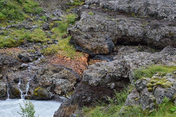 A close-up view of the rugged Icelandic terrain, featuring layers of dark volcanic rock, orange mineral staining, and a small stream of water. The image showcases the complex geological textures and diverse plant life found in a lava field environment.