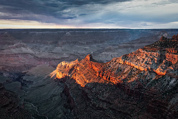 Sunlight illuminates canyon cliffs beneath dark storm clouds at the Grand Canyon. Iconic panoramic view from Pipe Creek Vista, showcasing the depth and geological layers of the vast landscape. Grand Canyon National Park, Arizona, United States, North America