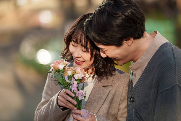Young man and woman lean close together while admiring a small bouquet of flowers, sharing a quiet and joyful moment. The woman smiles softly as she holds the flowers, while her partner looks on with warmth and affection. Gentle natural light and a softly blurred background create an intimate atmosphere that highlights emotional connection, tenderness and shared happiness. Ideal for concepts of romance, dating, Valentine’s Day, relationships, appreciation and meaningful everyday moments.