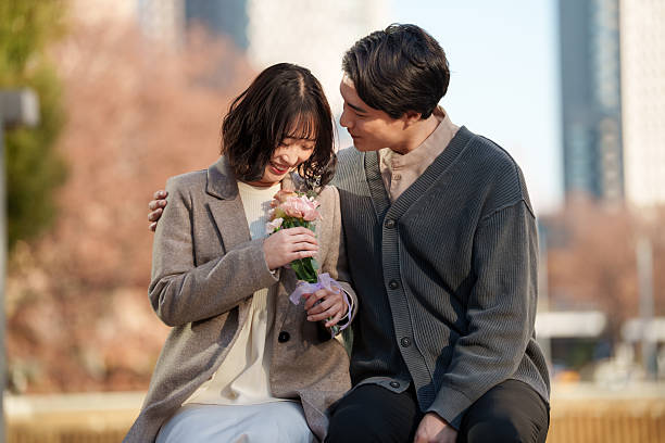 Young man and woman lean close together while admiring a small bouquet of flowers, sharing a quiet and joyful moment. The woman smiles softly as she holds the flowers, while her partner looks on with warmth and affection. Gentle natural light and a softly blurred background create an intimate atmosphere that highlights emotional connection, tenderness and shared happiness. Ideal for concepts of romance, dating, Valentine’s Day, relationships, appreciation and meaningful everyday moments.