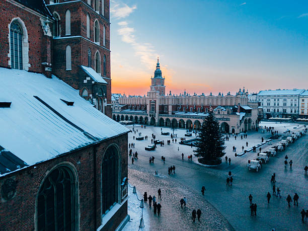 Aerial view of Krakow's historic Main Market Square (Rynek Główny) at sunset in winter. Snow-covered rooftops surround the square as warm orange and pink hues fill the sky. The iconic towers of St. Mary's Basilica and the Town Hall Tower rise above the old town, capturing the timeless beauty, architectural heritage, and romantic atmosphere of Krakow, Poland.