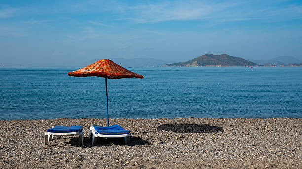 Quiet pebble beach with two sunbeds and a straw umbrella by the sea. Summer vacation, relaxation, and off-season travel concept.