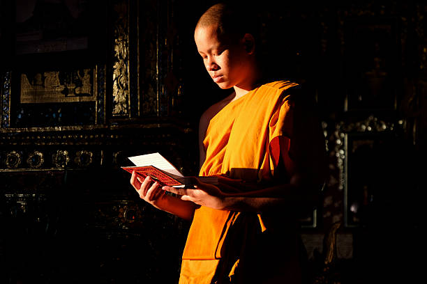 Buddhist novice wearing a robe reads a book by a window with soft morning light