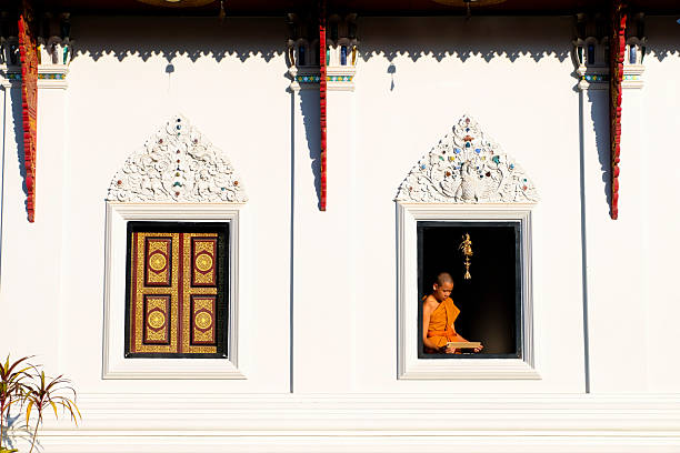 Buddhist novice wearing a robe reads a book by a window with soft morning light
