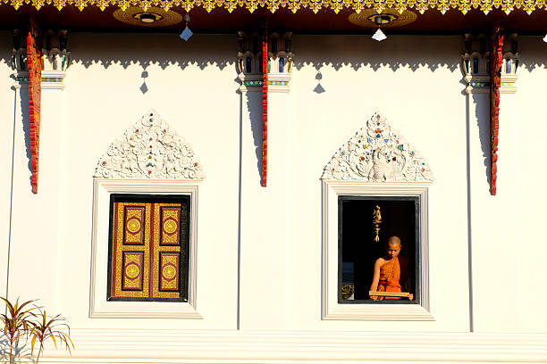 Buddhist novice wearing a robe reads a book by a window with soft morning light