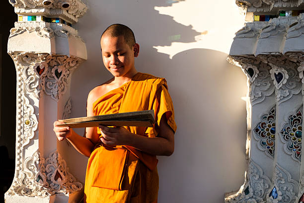 Buddhist novice wearing a robe reads a book by a window with soft morning light