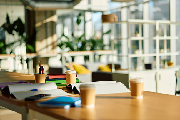 Unoccupied office desk inviting collaboration and productivity with open notebooks, pens, and paper coffee cups, creating a bright and modern workspace in a blurred corporate setting
