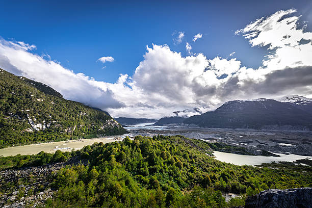 Wide-angle scene of a winding river flowing through green hills and steep mountain slopes near the Exploradores Glacier. The image represents untouched Patagonian wilderness and adventure travel in the Aysén region.