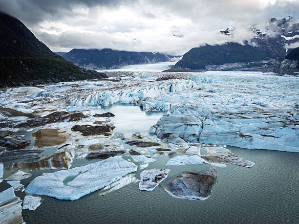 Close view of broken ice blocks drifting across a pale blue glacial lagoon beneath towering mountain walls. The image captures the fragile, constantly changing nature of the Exploradores Glacier environment.