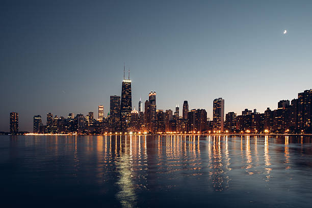 Night view of an urban skyline illuminated by building lights and reflected on water. The scene represents city nightlife, architecture, and metropolitan atmosphere after dark.