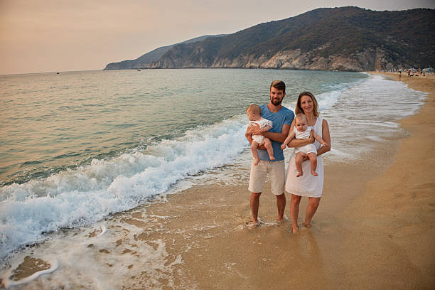 A family walks hand in hand down a tropical paradise beach during sunset. Mother and father are carrying baby twin girls.
