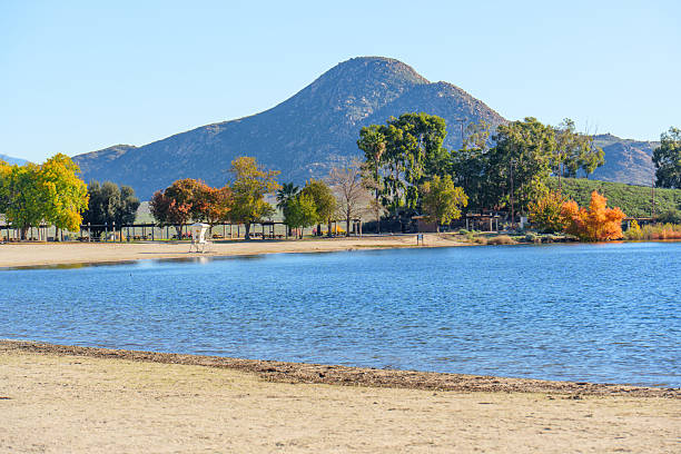 Lake Perris State Recreation Area's shoreline showcases vibrant autumn foliage against serene waters and rugged mountains. A lifeguard stand sits on the sandy beach.