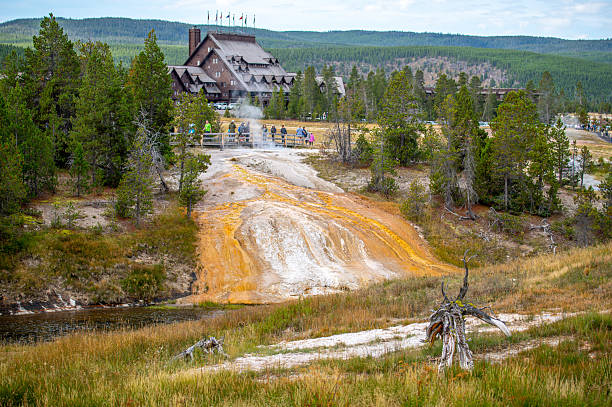 Geothermal feature flowing from beneath the boardwalk down a hillside into Yellowstone's Firehole River, with visitors observing and the historic lodge in the distance.