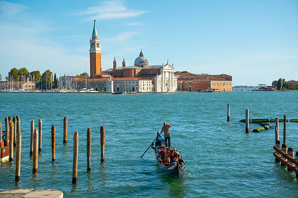 Venice, Italy - September 23, 2023: Gondolier standing and rowing a gondola with tourists on the venice lagoon, passing wooden poles with the iconic san giorgio maggiore island in the background