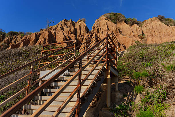 Rustic metal staircase ascends rugged sandstone cliffs at El Matador State Beach under a clear blue sky, revealing a dramatic coastal landscape.