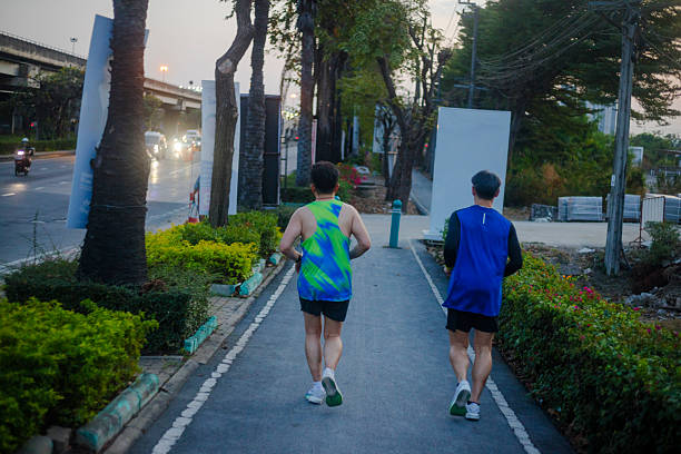 Two mature thai men are jogging in sunset along bicycle lane and sidealk of Pradit manutham Road  in  Bangkok Ladprao. Rear view of men wearing sports clothing. Scene is near THE WALK. At left side is traffic towards north and Ramintra