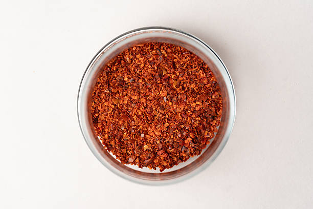 A high-angle, close-up shot of vibrant red chili flakes (pul biber) inside a clear glass jar. The image showcases the rich texture of crushed dried peppers and seeds, presented against a clean white background. Ideal for culinary blogs, spice shop marketing, ethnic food packaging, and spicy cuisine themes.