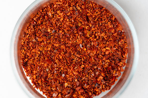 A high-angle, close-up shot of vibrant red chili flakes (pul biber) inside a clear glass jar. The image showcases the rich texture of crushed dried peppers and seeds, presented against a clean white background. Ideal for culinary blogs, spice shop marketing, ethnic food packaging, and spicy cuisine themes.