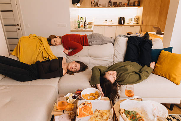 A cozy home scene shows three friends napping on a sectional after a casual pizza night. Soft lighting, blankets, and snacks create a warm, relaxed mood of friendship and comfort.