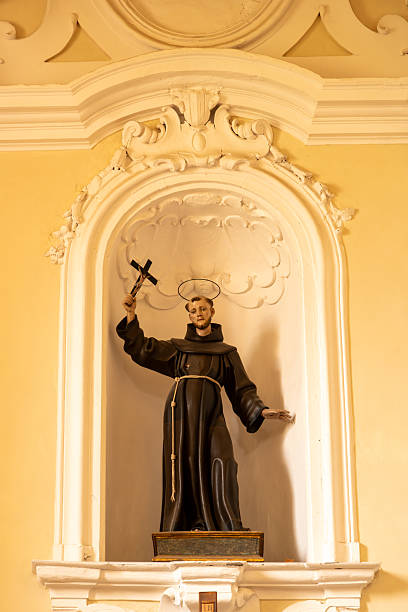 Sessa Aurunca â Campania â Italy â 08-29-2025: A robed monk statue with halo and raised cross stands in a white arched church niche against a warm yellow wall, lit by soft interior light.