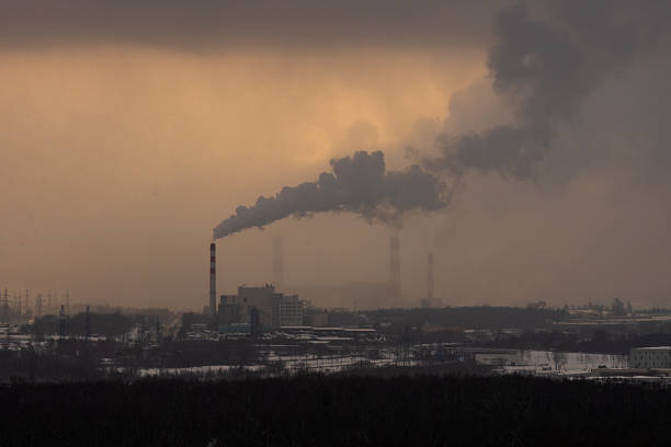Wide horizontal view of power plant chimneys emitting heavy smoke into a dense, muted atmosphere with low visibility above an industrial urban background. Image illustrates air pollution and industrial emissions with strong haze and available copy space.