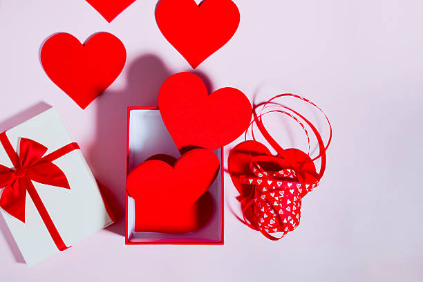 Open red gift box filled with heart shapes, styled with red paper hearts and a curled heart-pattern ribbon on a pink background. Playful, loving Valentine’s Day or Mother’s Day layout with copy space for messaging.