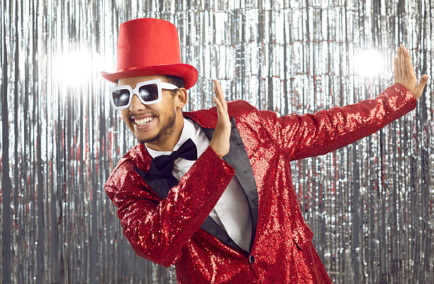 Young positive african american showman in shiny suit having fun on silver background with sparkle decoration. Joyful guy in red disco suit, hat and sunglasses smiles broadly at camera while dancing.